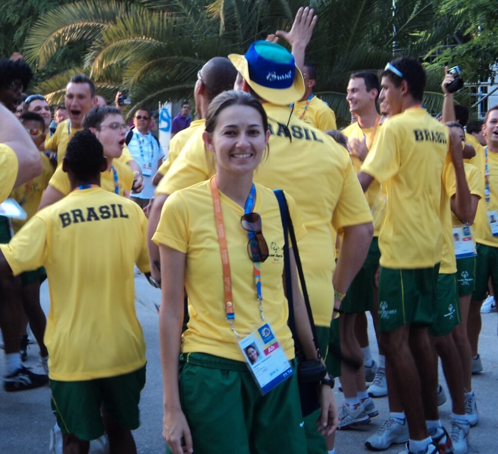 Descrição da imagem #pracegover: Foto de Thais em um evento internacional. Ela é uma mulher branca de cabelos castanhos que estão presos. Thais veste uma camiseta amarela e calça verde do uniforme. Ao fundo vemos outras pessoas, entre membros da comissão técnica e atletas, que também estão com camiseta amarela e calça verde. Fim da descrição.