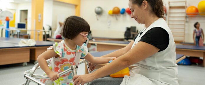 Descrição da imagem #pracegover: Foto de uma criança em atendimento com uma profissional da AACD. A criança é uma menina branca de cabelos castanhos lisos e curtos. Ela usa um óculos rosa e está vestindo uma blusa colorida. A menina usa um andador e está sendo auxiliada por uma mulher branca de cabelos castanhos que estão amarrados. A mulher está vestindo uma blusa preta em baixo de um jaleco branco. Ao fundo vemos objetos da sala de reabilitação da AACD. Fim da descrição.