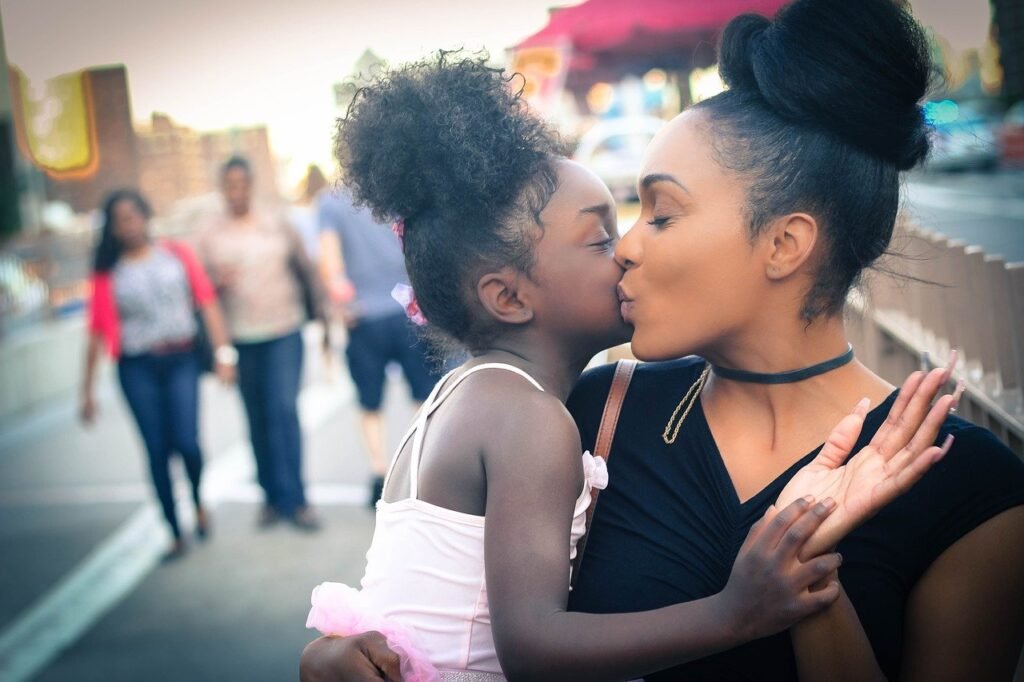 Descrição da imagem #pracegover: Foto de uma mulher beijando uma menina que está em seu colo. A mulher e a menina são negras e estão com o cabelo preso em um coque alto. A mulher usa blusa preta e a menina uma blusa rosa. Ao fundo vemos pessoas que passam pelo local. Fim da descrição.