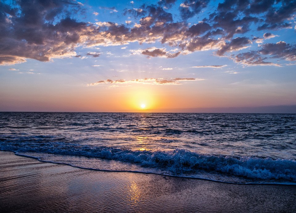Descrição da imagem #pracegover: Foto do amanhecer na praia. Na parte inferior está a faixa de areia molhada pelas ondas do mar. Ao fundo o sol está nascendo e na parte superior estão algumas nuvens.