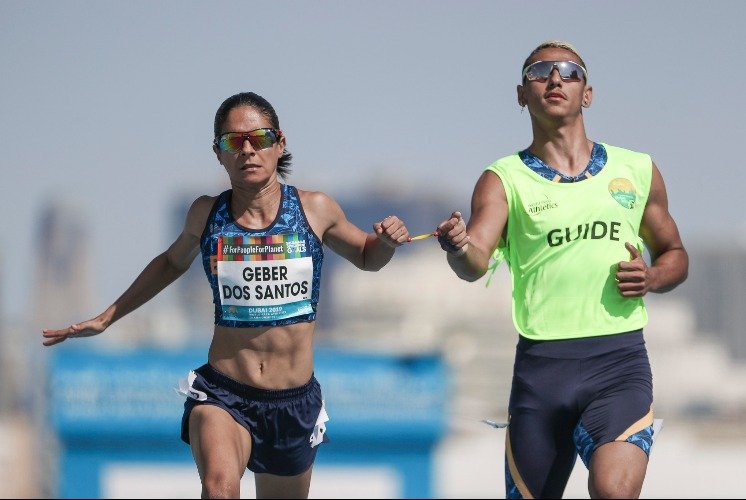 Descrição da imagem #pracegover: A paratleta Jerusa Geber durante uma de suas disputas. Jerusa é uma mulher branca com cabelos castanhos que estão amarrados. Ela tem deficiência visual e está correndo acompanhada de seu guia. Fim da descrição.