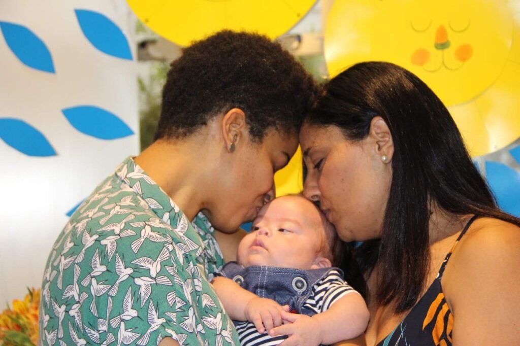 Descrição da imagem #pracegover: Foto de duas mulheres beijando um bebê com síndrome de Down. Fim da descrição.