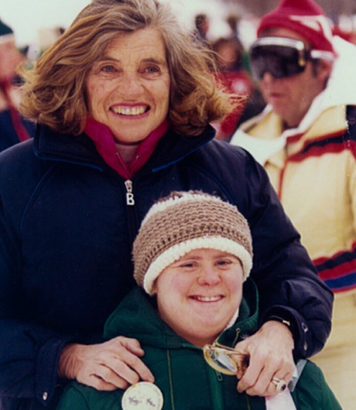 Descrição da imagem #pracegover: Foto de Eunice Kennedy Shriver e uma jovem atleta da Special Olympics. Eunice está em pé, atrás da atleta. Eunice é uma mulher branca, de cabelos loiros na altura dos ombros. Ela está vestindo um casaco azul marinho e um lenço vinho no pescoço. A atleta tem síndrome de Down. Ela é uma mulher branca, que está sentada. Ela veste um casaco verde e usa um gorro de lã na cabeça. As duas estão sorrindo. Ao fundo estão algumas pessoas com roupas de frio. Fim da descrição.