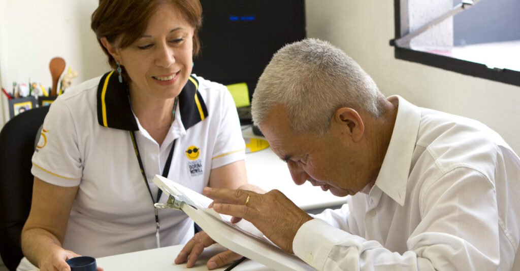 Descrição da imagem #pracegover: Foto de um atendimento na Fundação Dorina. Um idoso está fazendo a leitura em um tablet sobre a mesa. Uma mulher, da equipe da Fundação, está ao seu lado. Fim da descrição.