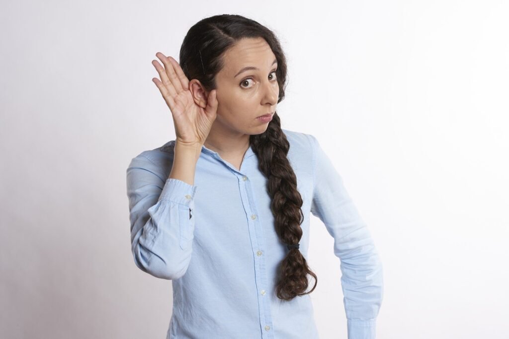 Descrição da imagem #pracegover: Uma mulher está com a mão direita na orelha e faz cara de quem está questionando a audição. A mulher é branca, tem cabelos castanhos compridos que estão em uma longa trança lateral. Ela veste uma camisa azul claro. Fim da descrição.