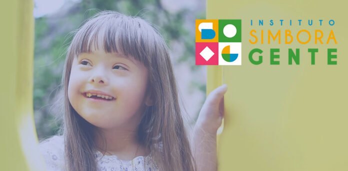 Descrição da imagem #pracegover: Imagem de uma menina com síndrome de Down. Ela está olhando para o lado e sorri. A menina é branca e tem cabelos castanhos, lisos e compridos. No canto superior direito está o logo do Instituto Simbora Gente, formado por imagens construídas a partir de figuras geométricas. Fim da descrição.
