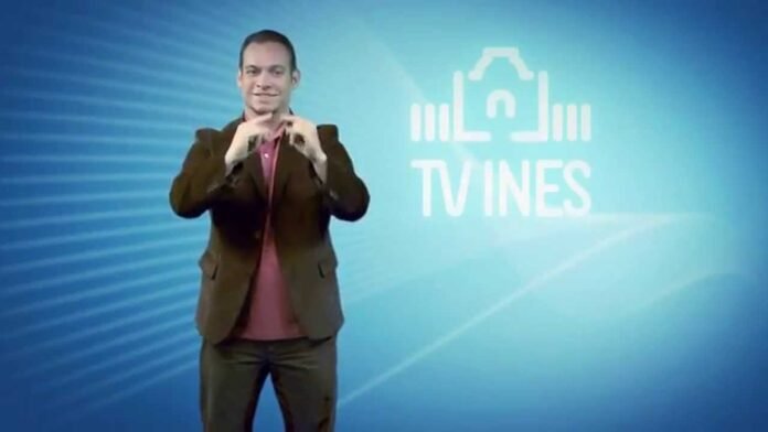Descrição da imagem #pracegover: Print de tela de um programa da TV INES. Um homem está de pé. Ele é branco, tem cabelos castanhos curtos e está vestindo um paletó marrom e camisa bordô. O homem está fazendo um sinal em Libras. Ao fundo vemos um painel azul com o logo da TV INES. Fim da descrição.