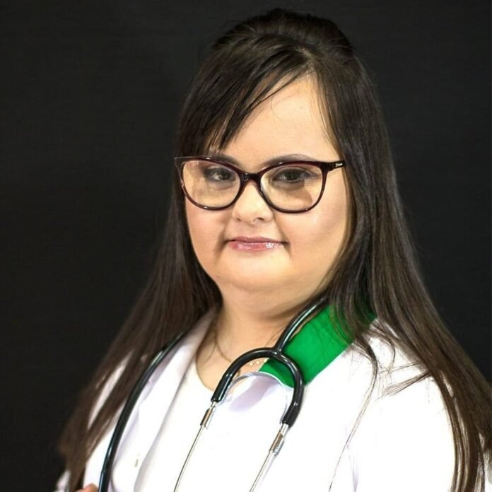Descrição da imagem #pracegover: Foto de Luana. Ela é uma mulher branca de cabelos pretos, lisos e compridos. Luana usa óculos. Ela tem síndrome de Down e está vestindo um jaleco branco e tem um estetoscópio sobre os ombros. Fim da descrição.