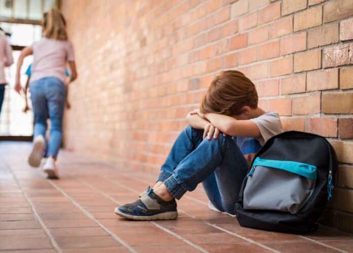 Descrição da imagem #pracegover: Um menino está sentado no chão e tem a cabeça abaixada sobre os braços. Ele está em uma escola. Fim da descrição.