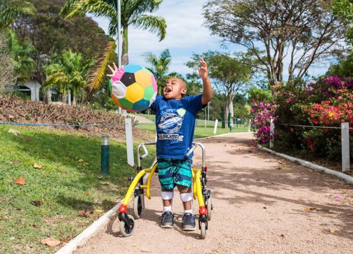 Descrição da imagem: Foto do menino João Gabriel, que tem paralisia cerebral. Ele usa órteses e um andador e está brincando com uma bola. Fim da descrição.
