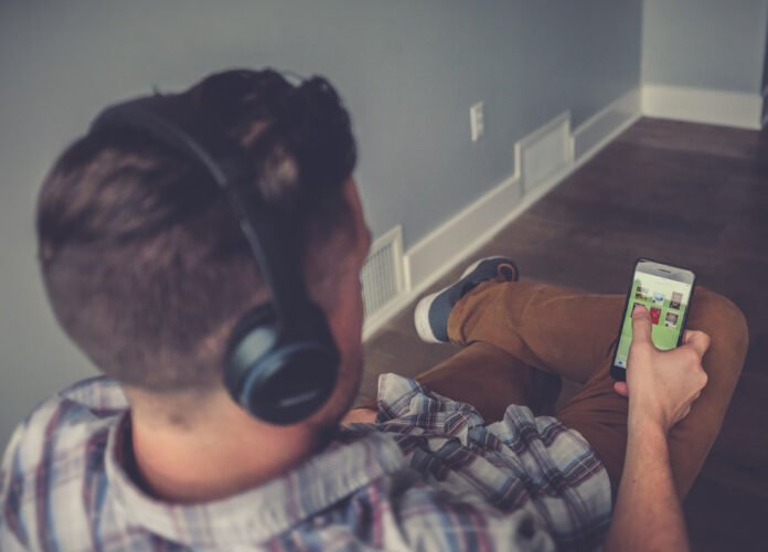 Descrição da imagem #pracegover: Foto de um rapaz ouvindo o audiolivro. Ele está sentado. Está usando um fone de ouvidos e segura o celular. Fim da descrição.
