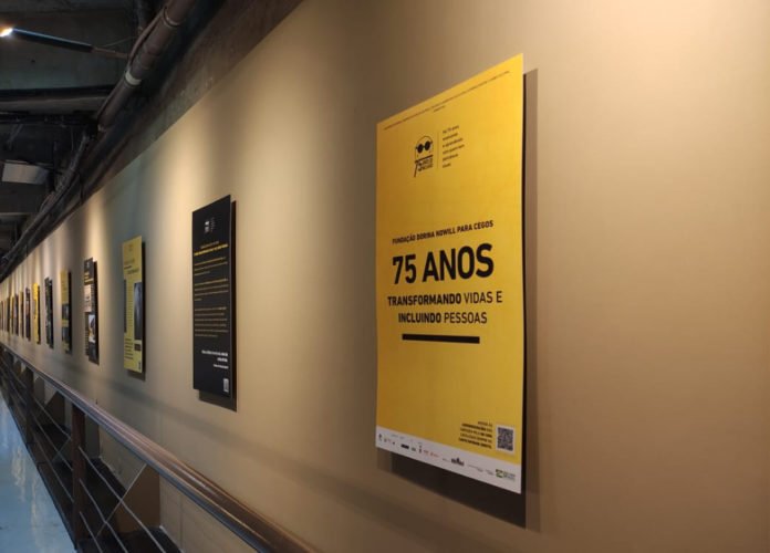 Descrição da imagem #pracegover: Foto do corredor da Unibes Cultural com a exposição sobre a história da Fundação Dorina. Vemos ao longo de um grande corredor alguns quadros. Fim da descrição.
