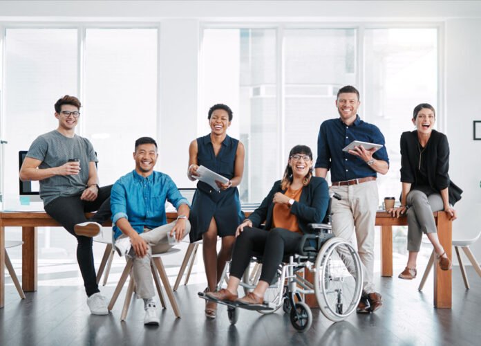 Descrição da imagem #pracegover: Foto de várias pessoas em um ambiente corporativo. Entre eles está uma jovem com deficiência física. Fim da descrição.