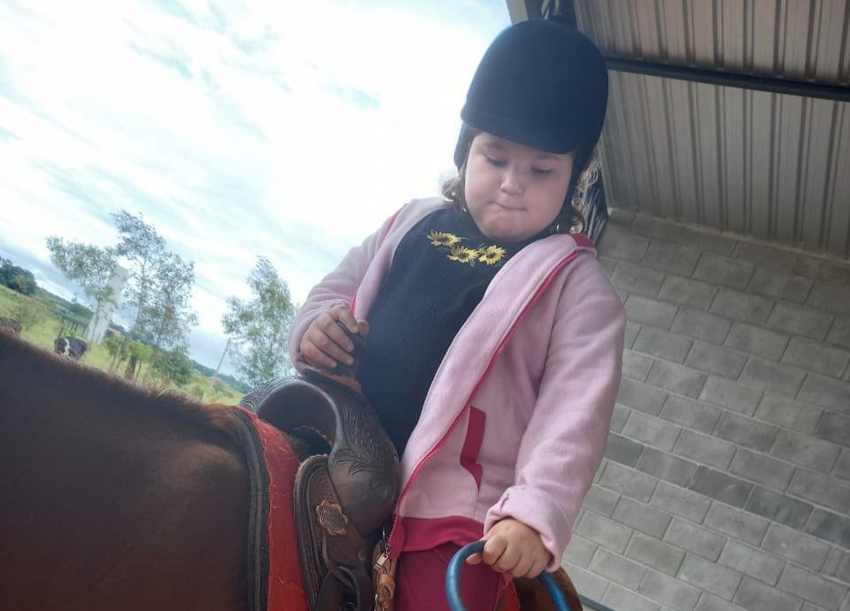 Descrição da imagem: Foto da menina Ester. Ela está montada em um cavalo marrom. Fim da descrição.