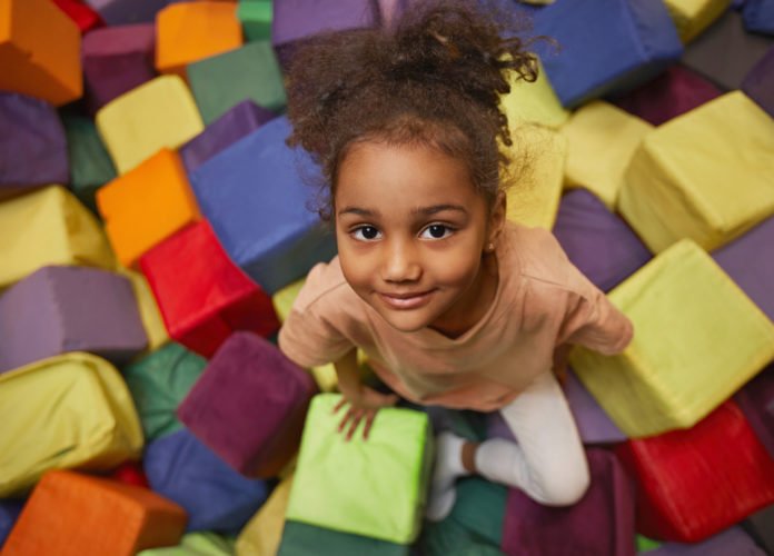 Descrição da imagem #pracegover: Foto de uma menina brincando sentada em meio a blocos coloridos de espuma. Fim da descrição.