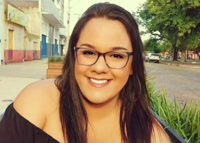 Descrição da imagem: Foto da Pietra Simon. Ela é uma mulher branca, de cabelos castanhos, lisos e compridos. Pietra está usando óculos de grau e sorri. Fim da descrição.