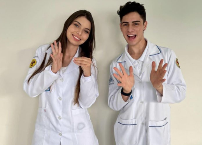 Descrição da imagem: Foto dos estudantes fazendo sinais em Libras. Eles estão usando jalecos brancos. Fim da descrição.