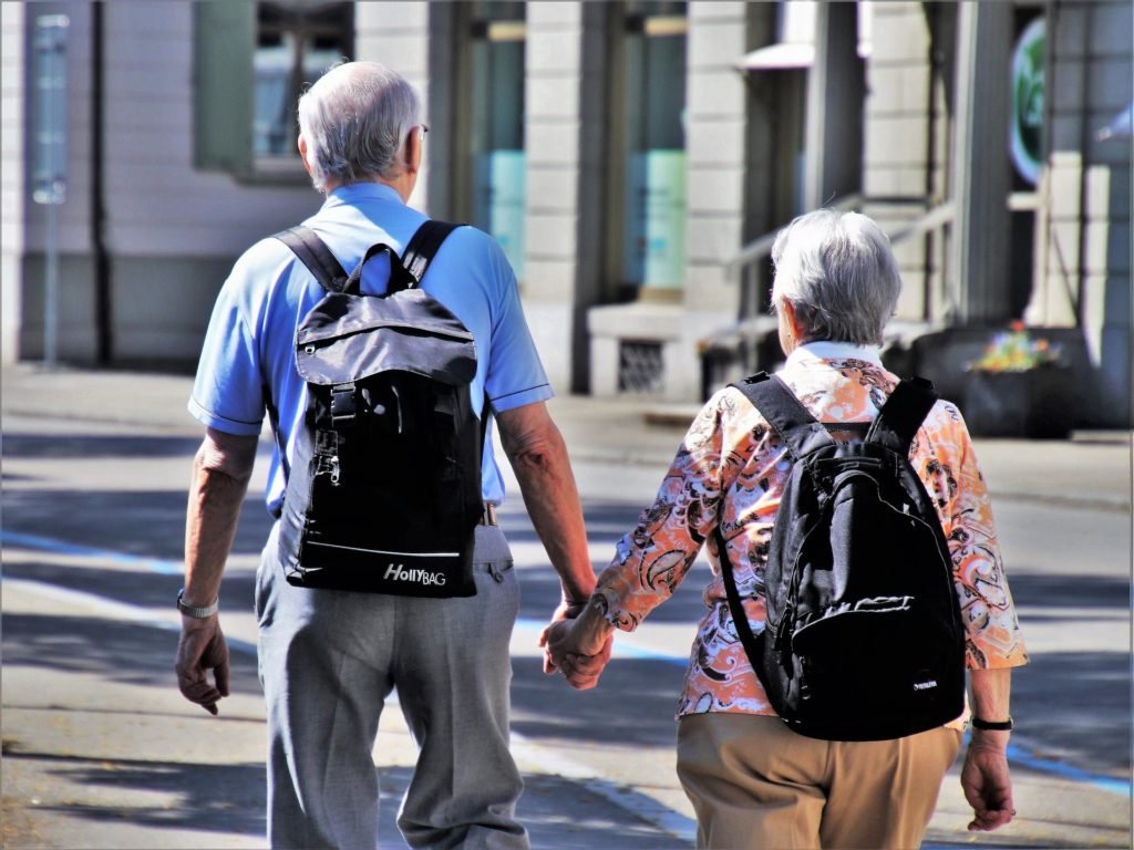 Descrição da imagem #pratodosverem: Um casal de idosos está andando em uma rua, de mãos dadas. Eles estão com mochilas nas costas. Fim da descrição.