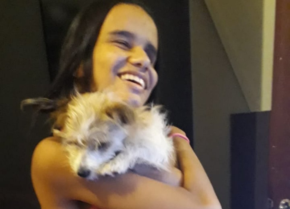 Descrição da imagem: Foto da Thaís Vitória com sua cadelinha Kyara. Thaís é jovem, tem cabelos castanhos. Ela está abraçando a cachorrinha e sorri. Fim da descrição.