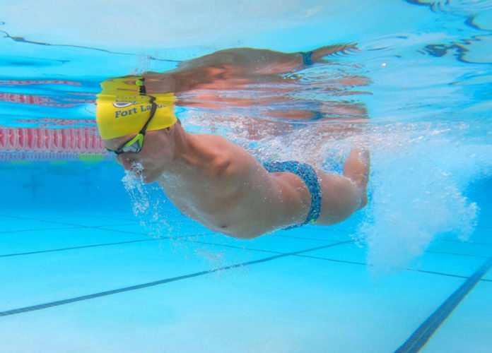 Descrição da imagem: Foto feita dentro de uma piscina do nadador Abbas Karimi. Ele está submerso, usa toca amarela e óculos de natação. Fim da descrição.