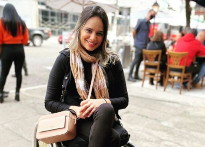 Descrição da imagem: Foto de Pamela. Ela está em sua cadeira de rodas. Pamela é um mulher loira, de cabelos lisos na altura dos ombros. Ela usa roupas pretas, cachecol e bolsa bege. Fim da descrição.