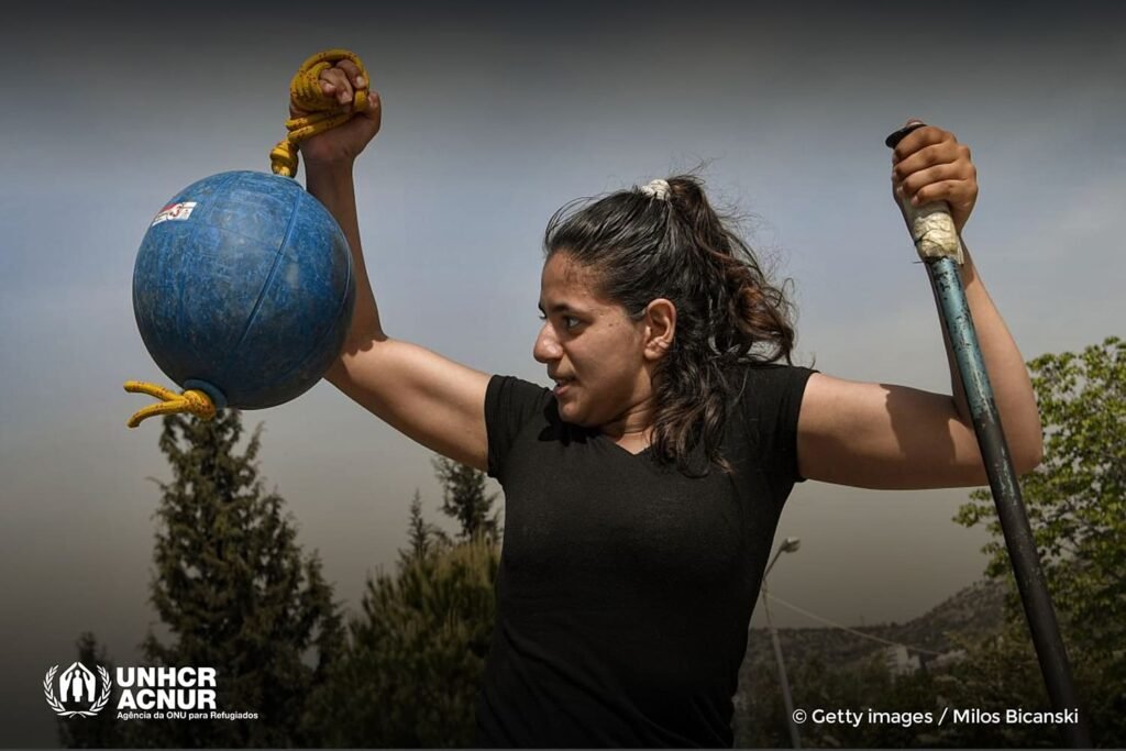 Descrição da imagem: Foto de Alia Issa. Ela está segurando um dardo e uma clava, utilizados na modalidade de paratletismo. Ela tem cabelos pretos e pele clara. Usa camiseta preta. Fim da descrição.
