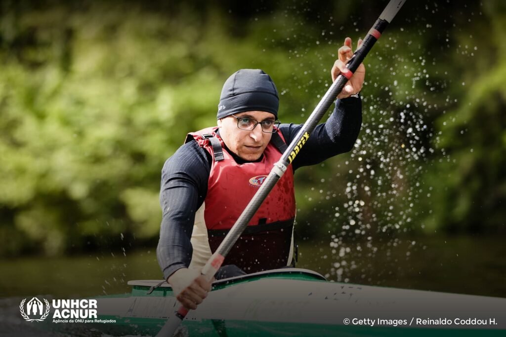 Descrição da imagem: Anas Al Khalifa está praticando canoagem. Tem a pele parda, usa óculos de grau, toca e camiseta preta, e colete salva-vidas vermelho. Fim da descrição.