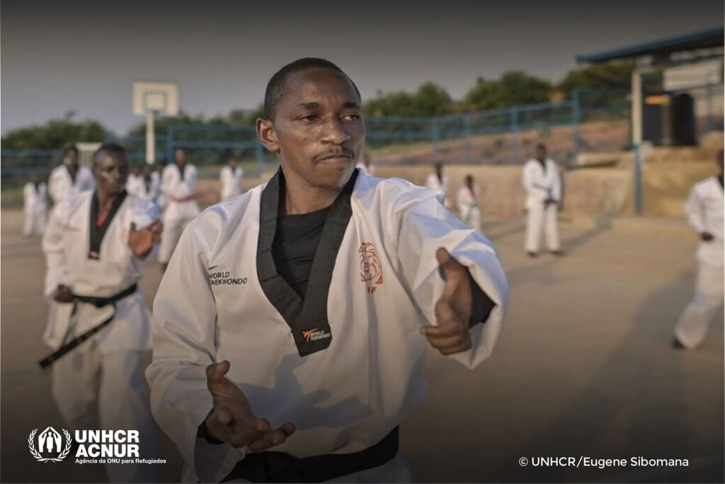 Descrição da imagem: Parfait Hakizimana está treinando Taekwondo em uma quadra descoberta, com outros atletas, em segundo plano. Ele tem pele negra, cabelos pretos curtos e usa kimono branco. Fim da descrição.