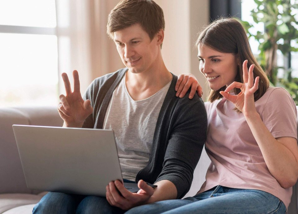 Descrição da imagem: Foto de um rapaz e de uma moça. Eles estão sentados e usam o computador para se comunicar. Ambos estão fazendo sinais com as mãos. Fim da descrição.