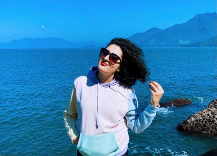 Descrição da imagem: Foto de Julyana Maia. Ela é uma moça de pele branca e cabelos pretos, cacheados, na altura dos ombros. Julyana está em uma praia. Atrás dela está o mar. Juliana está vestindo um moleton rosa com as mangas azuis. Fim da descrição.