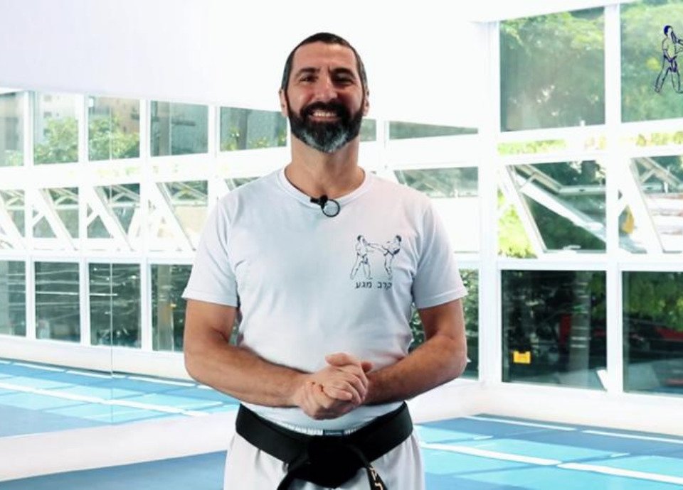 Descrição da imagem: Foto de Avigdor Zalmon. Ele é um homem branco de barba e cabelos grisalhos. Ele está num local de treino e usa calça e camiseta brancas e uma faixa preta na cintura. Fim da descrição.