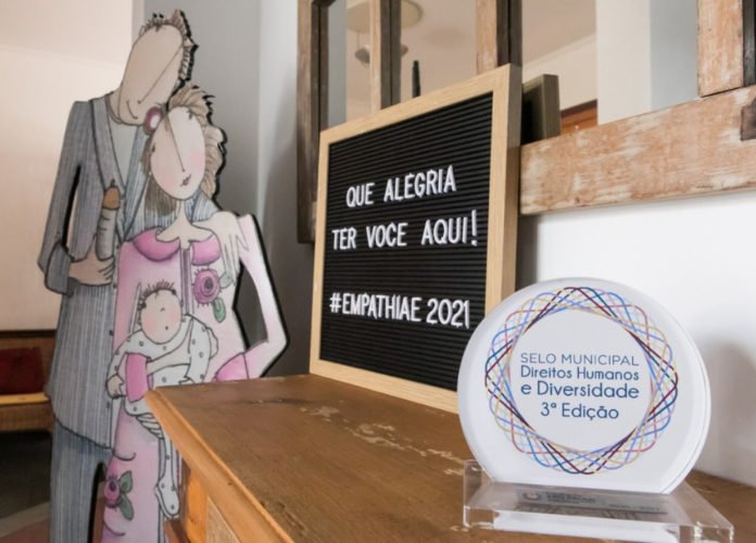 Descrição da imagem: Foto de alguns detalhes da sede do Instituto Empathiae. Sobre um balcão de madeira estão um quadrinho, onde está escrito: 'Que alegria ter você aqui' e uma placa do 'Selo Municipal Direitos Humanos e Diversidade'. Fim da descrição.