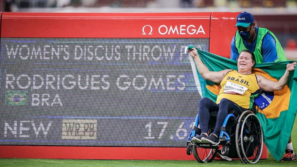 Descrição da imagem: Foto da atleta Beth Gomes comemorando suas conquists. Ela está ao lado do painel onde está registrado seu recorde mundial. Fim da descrição.