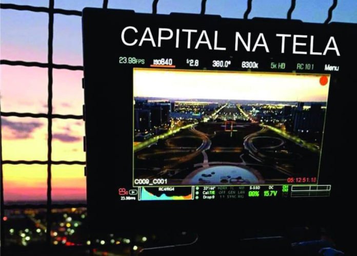 Descrição da imagem: Foto de um cartaz da mostra Capital na Tela. Ao fundo está uma janela com um lindo por do sol. Fim da descrição.