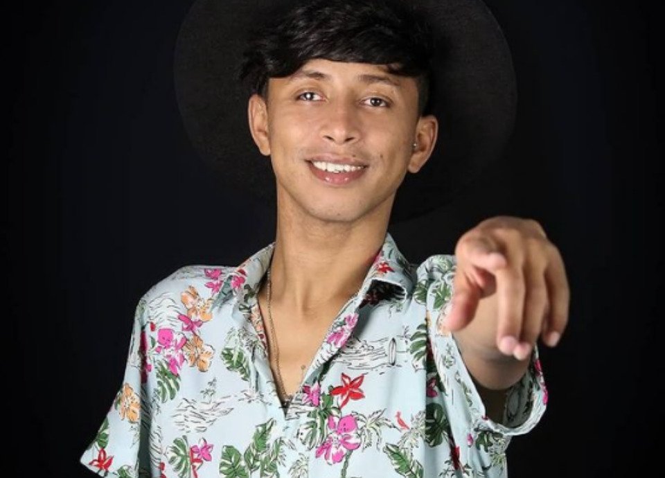 Descrição da imagem: Foto de Daniel Massafera. Ele está com o braço esquerdo esticado, apontando para a frente. Daniel é um rapaz de cabelos pretos, lisos e curtos. Ele está vestindo uma camisa de manga curta florida. Fim da descrição.