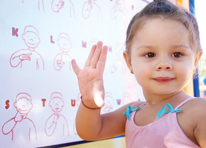 Descrição da imagem: Foto de uma menina. Ela está fazendo um sinal em Libras. Atrás dela vemos um painel com os sinais das letras em Libras. Fim da descrição.