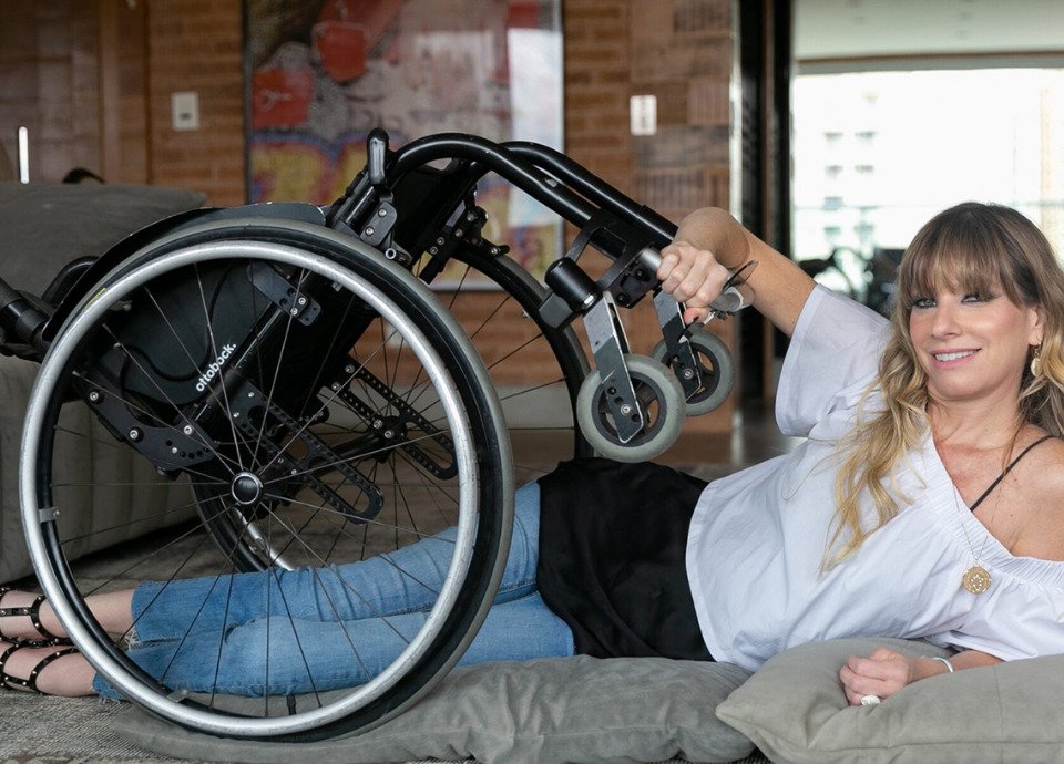 Descrição da imagem: Foto de Andrea Schwarz. Ela é uma mulher branca e tem cabelos longos, loiros. Andréa está deitada no chão, de lado, e está segudando sua cadeira sobre seu corpo. Fim da descrição.