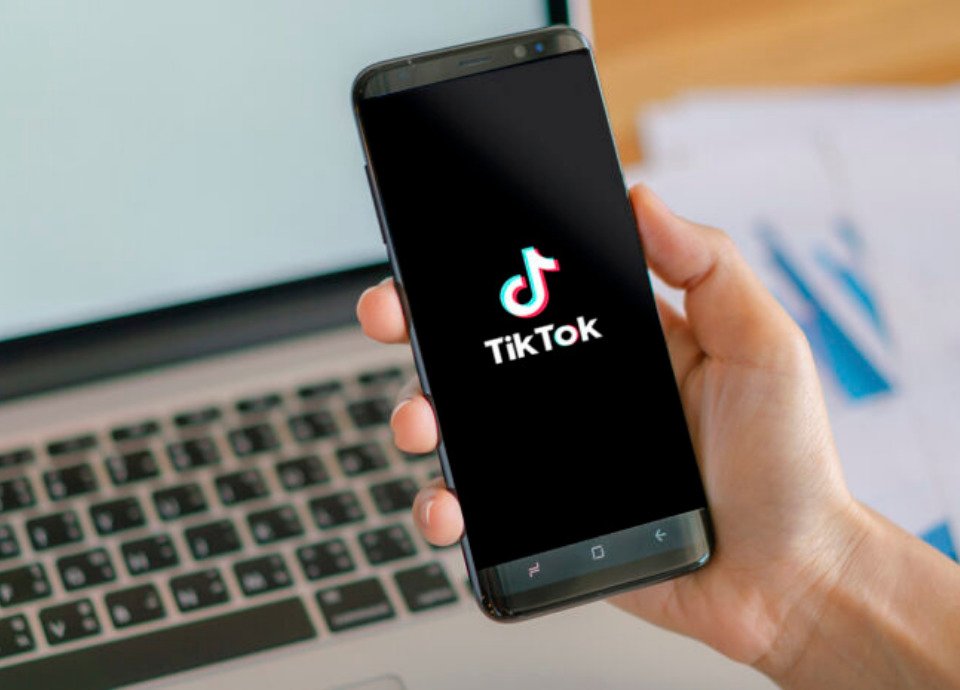 Descrição da imagem: Foto de uma pessoa segurando um celular com o aplicativo Tik Tok em destaque. Ao fundo está um computador. Fim da descrição.