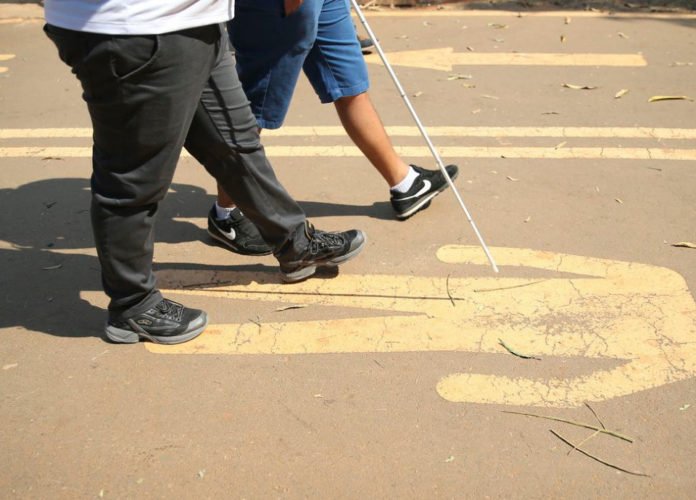 Descrição da imagem: Foto dos pés de uma pessoa com deficiência visual caminhando na rua. Ela está usando uma bengala e conta com a companhia de outra pessoa, que caminha ao seu lado. Fim da descrição.
