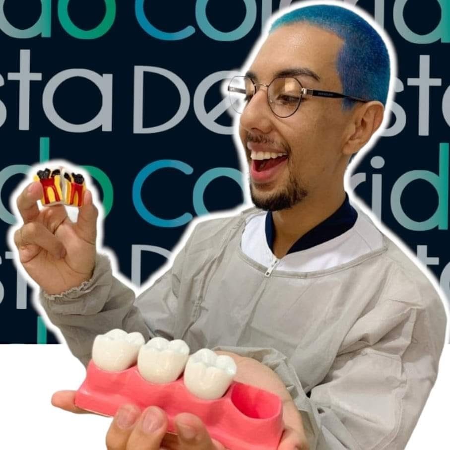 #descriçãodaimagem: Foto do Jefferson. Ele é um homem jovem, usa óculos de grau e tem os cabelos curtos, pintados de azul. Jefferson usa roupa de dentista e segura alguns objetos que remetem à odontologia. Fim da descrição.