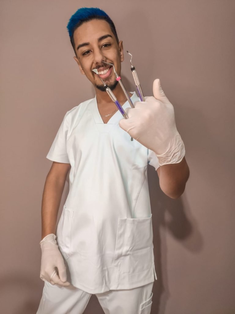 #descriçãodaimagem: Foto do Jefferson. Ele é um homem jovem e tem os cabelos curtos, pintados de azul. Jefferson usa roupa de dentista e segura, entre os dedos, alguns instrumentos de odontologia. Fim da descrição.