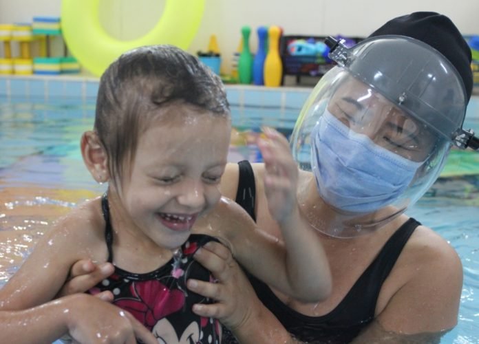 Descrição da imagem: Foto de uma menina durante atividade na piscina da AACD. A criança está acompanhada por uma profissional da AACD, que a segura. A menina usa maiô e está sorrindo. A profissional usa máscara e face shield. Fim da descrição.