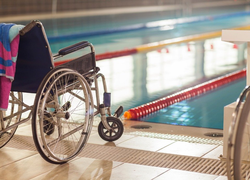 Descrição da imagem: Foto de uma cadeira de rodas na beira de uma piscina com raias olímpicas. Fim da descrição.