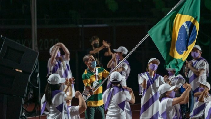 Descrição da imagem: Foto da cerimônia de encerramento dos Jogos Paralímpicos. O atleta Daniel Dias está em destaque, no centro da imagem. Ele está carregando a bandeira do Brasil. Fim da descrição.