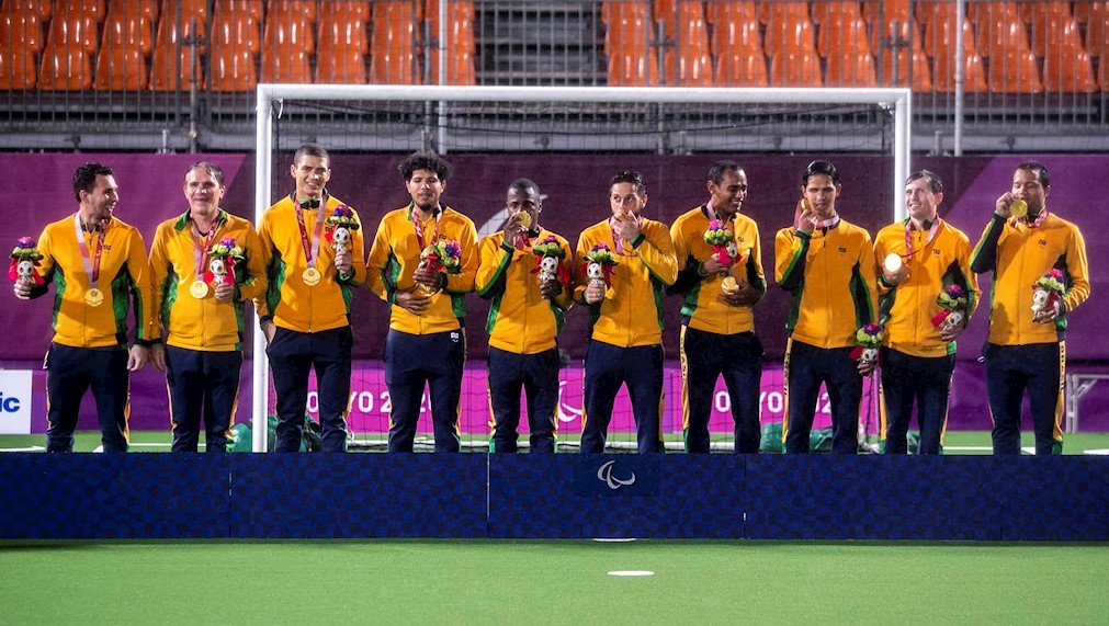 Descrição da imagem: Foto dos atletas da selecão de futebol de 5. Eles estão no pódio recebendo suas medalhas de ouro. Fim da descrição.