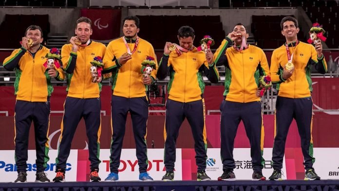 Descrição da imagem: Foto dos atletas da selecão de goalball no pódio recebendo suas medalhas de ouro. Fim da descrição.
