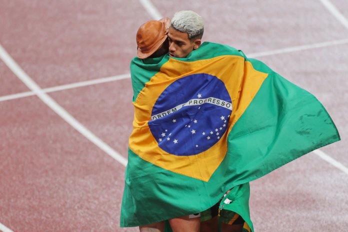 Descrição da imagem: Foto dos atletas Petrúcio Ferreira e Thomaz Moraes se abraçando após disputa no atletismo. Eles estão na pista de atletismo, logo após terem conquistado medalhas de prata e bronze. Eles estão enrolados na bandeira do Brasil e Petrúcio usa um chapeu de cangaceiro, em referência ao nordeste. Fim da descrição.