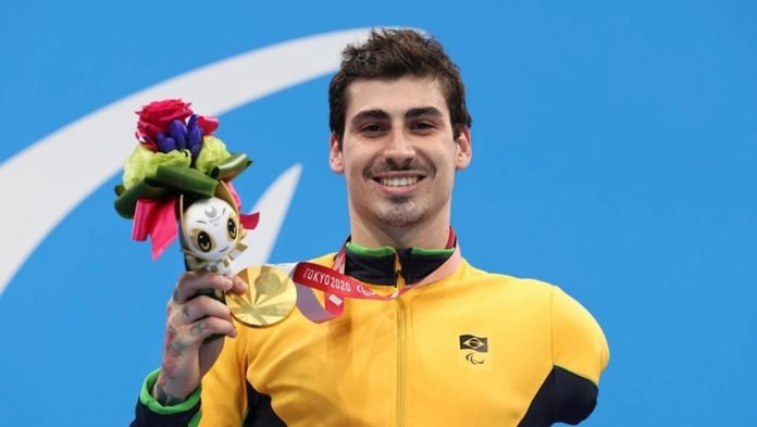 Descrição da imagem: Foto do atleta Talisson Glock no pódio com sua medalha de ouro. Talisson é um rapaz branco de cabelos castanhos, curtos. Ela está vestindo o uniforme amarelo da selecão. Fim da descrição.