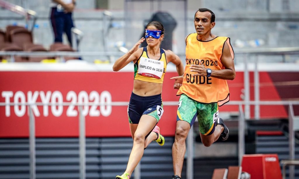 Descrição da imagem: Foto a atleta Thalita Simplicio. Ela está na pista, acompanhada de seu guia, durante competição de atletismo. Fim da descrição.   