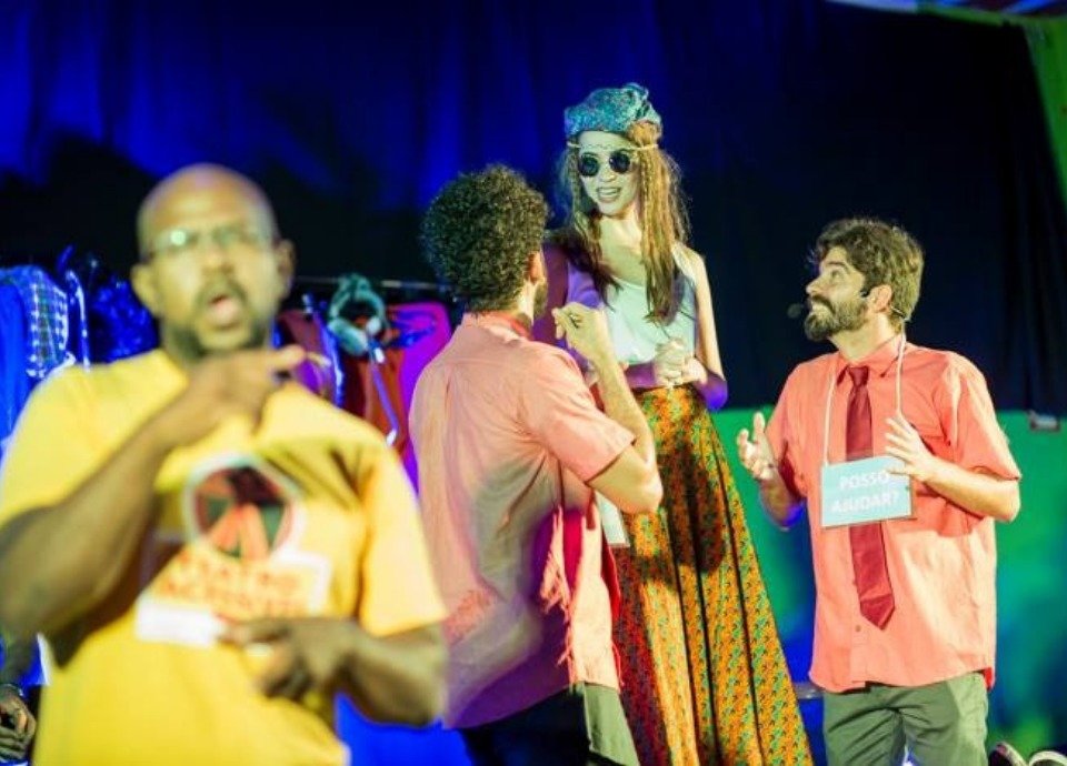 #descriçãodaimagem: Foto de uma apresentação teatral com interpretação em Libras. Ao fundo vemos os atores no palco e em primeiro plano, vemos o intérprete de Libras, traduzindo a apresentação. Fim da descrição.